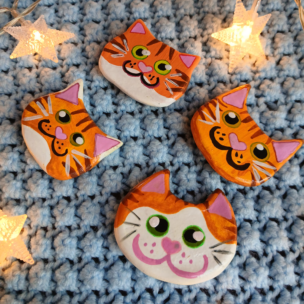 Four orange tabby cat face magnets all with different hand-painted faces arranged on a textured light blue background surrounded by star string lights.