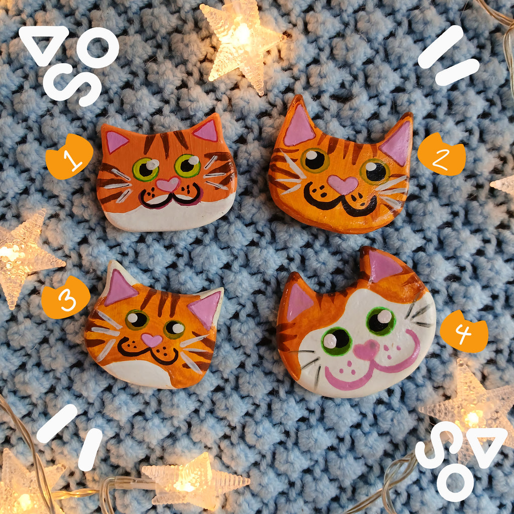 Four orange tabby cat face magnets all with different hand-painted faces arranged on a textured light blue background surrounded by star string lights.