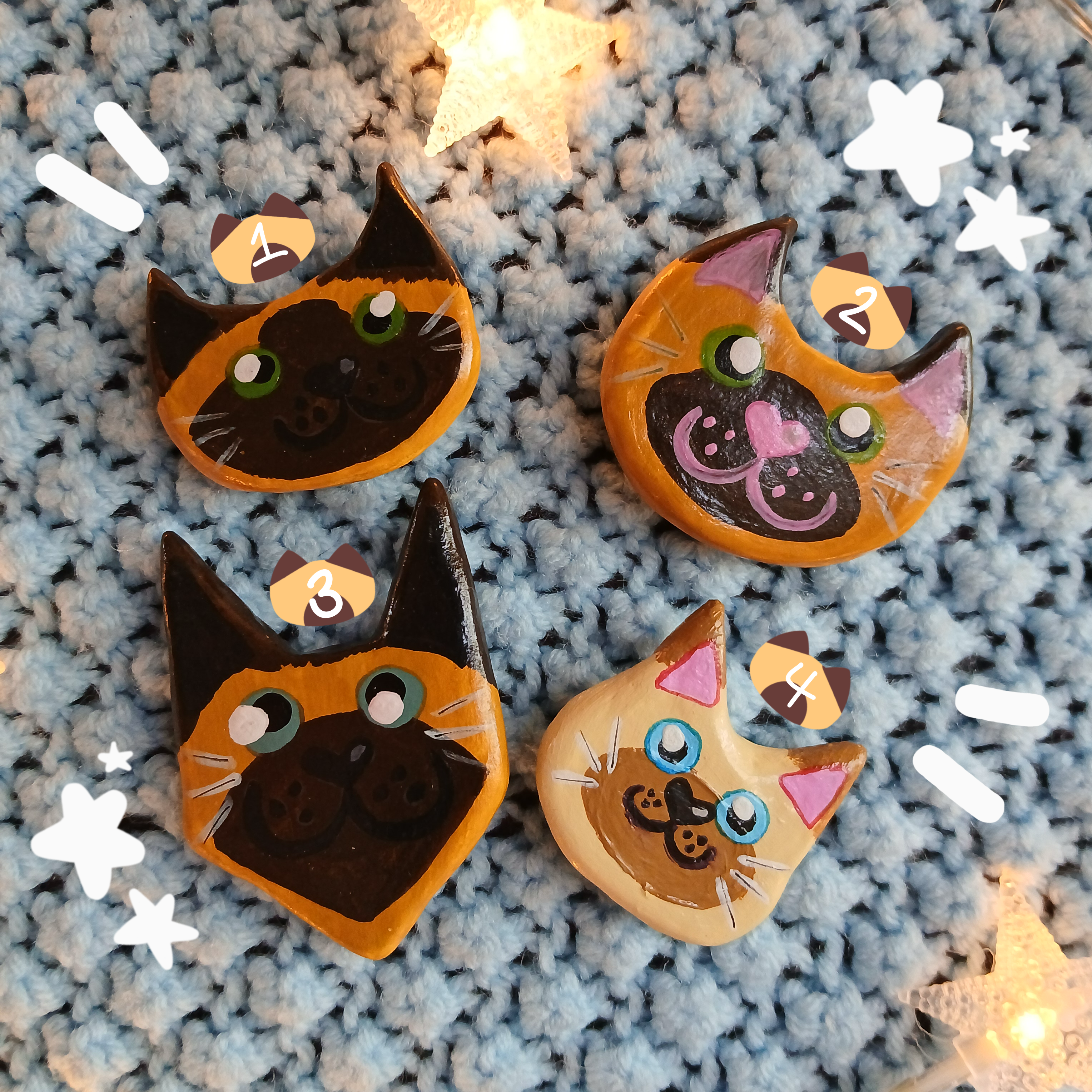 Four colorpoint cat face magnets all with different hand-painted faces arranged on a textured light blue background surrounded by star string lights.