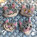 Four gray tabby cat face magnets all with different hand-painted faces arranged on a textured light blue background surrounded by star string lights.