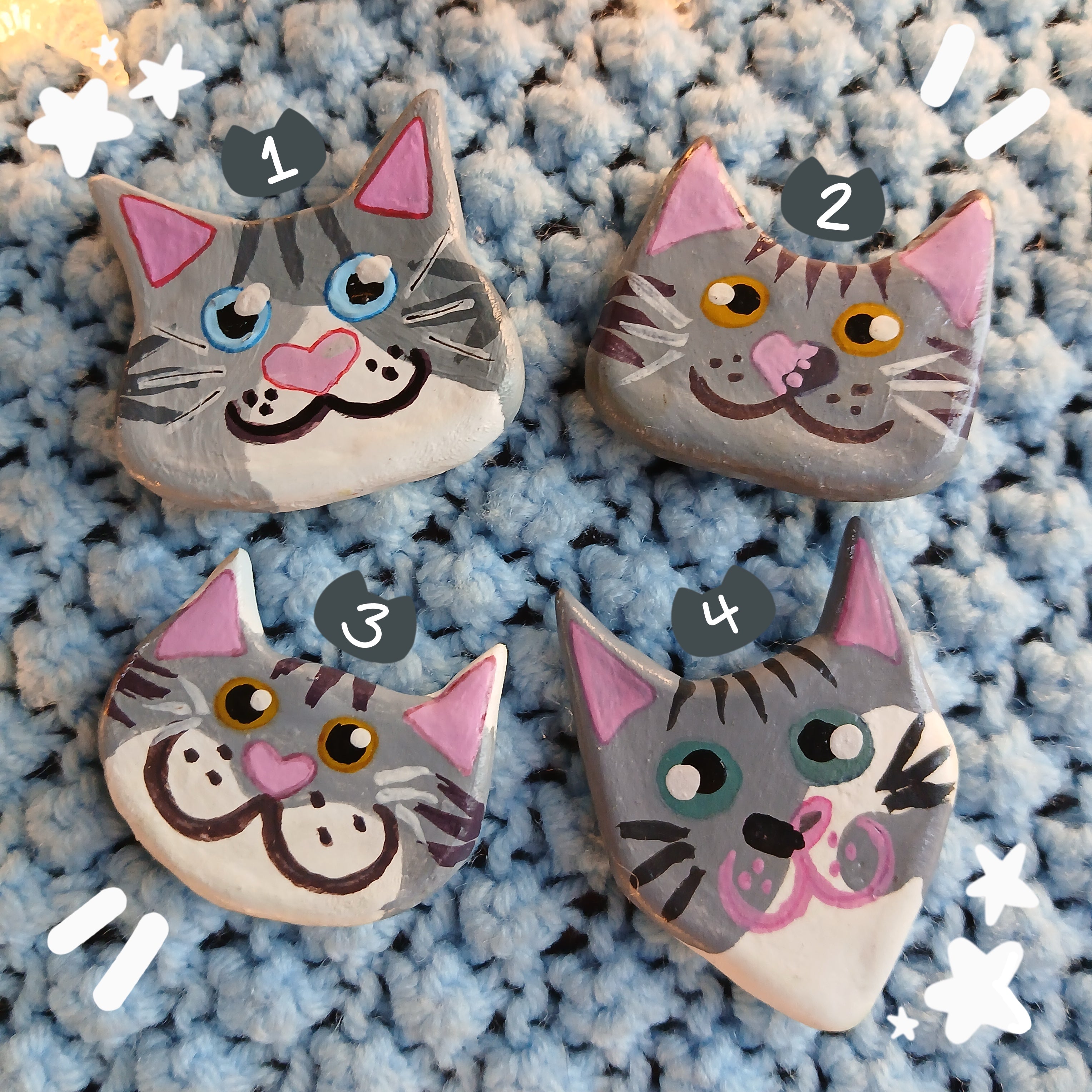 Four gray tabby cat face magnets all with different hand-painted faces arranged on a textured light blue background surrounded by star string lights.