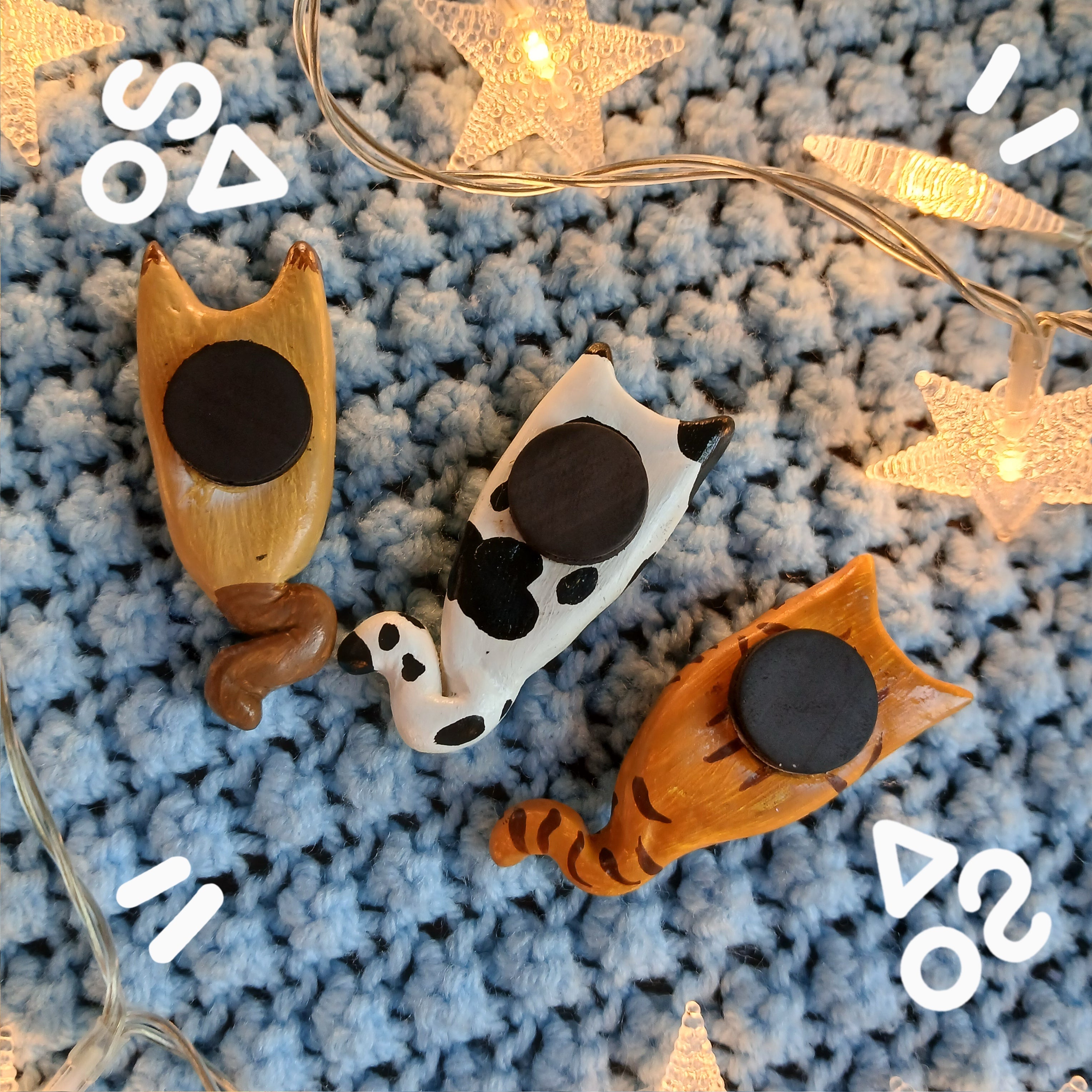 Collection of clay cat-shaped magnets, face down showing the magnets on the back, on light blue textured background, with decorative star lights.