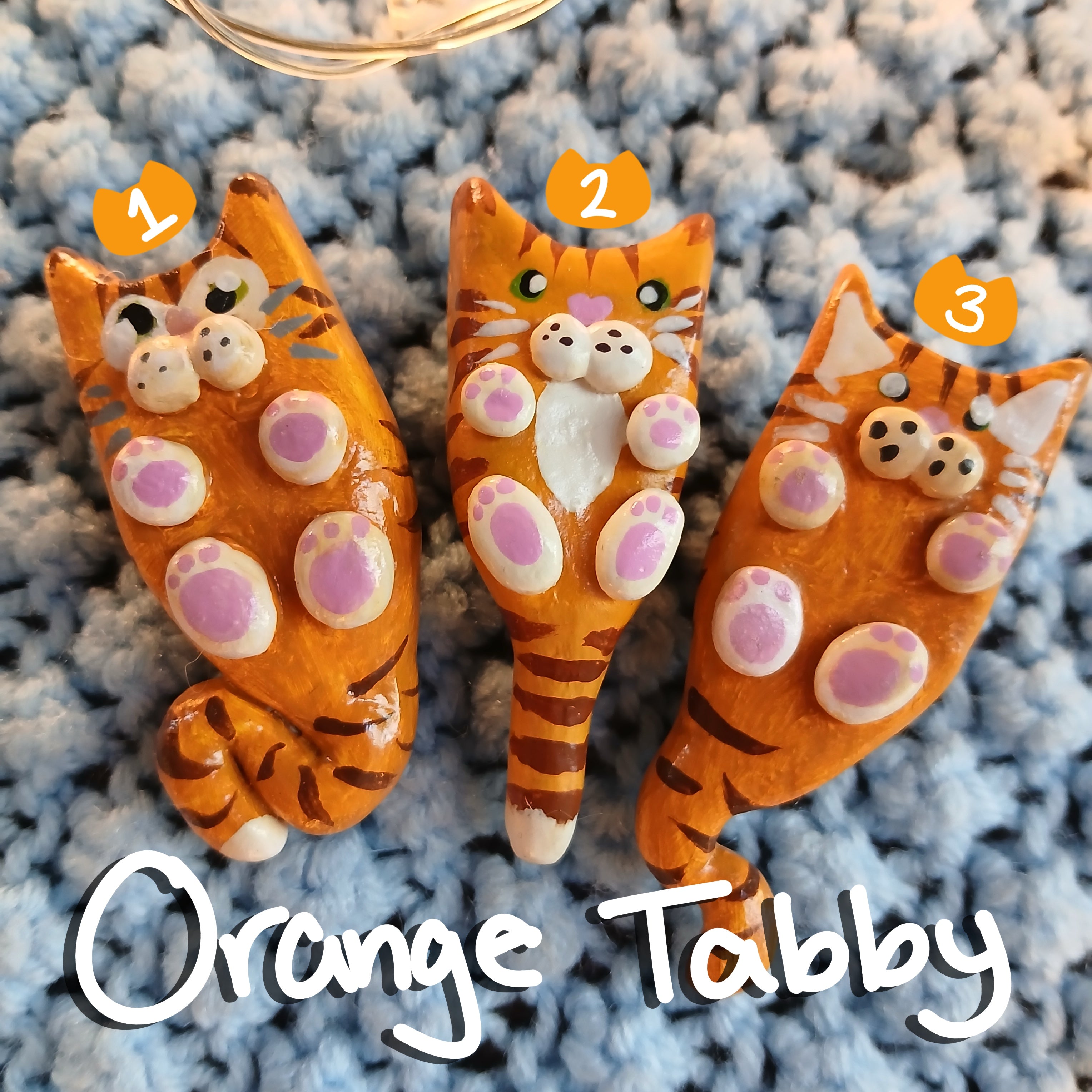 Three clay orange tabby cat-shaped magnets, on light blue textured background, with decorative star lights.