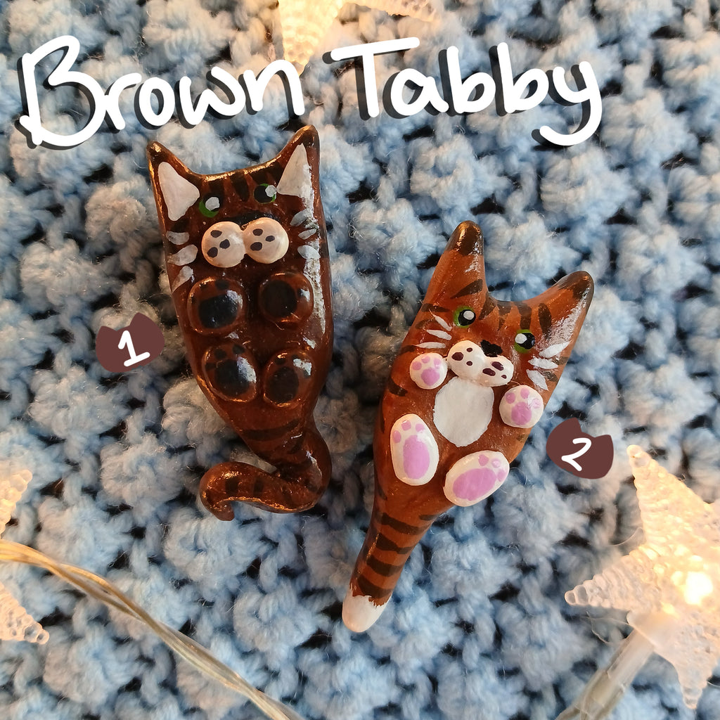 Two clay brown tabby cat-shaped magnets, on light blue textured background, with decorative star lights.