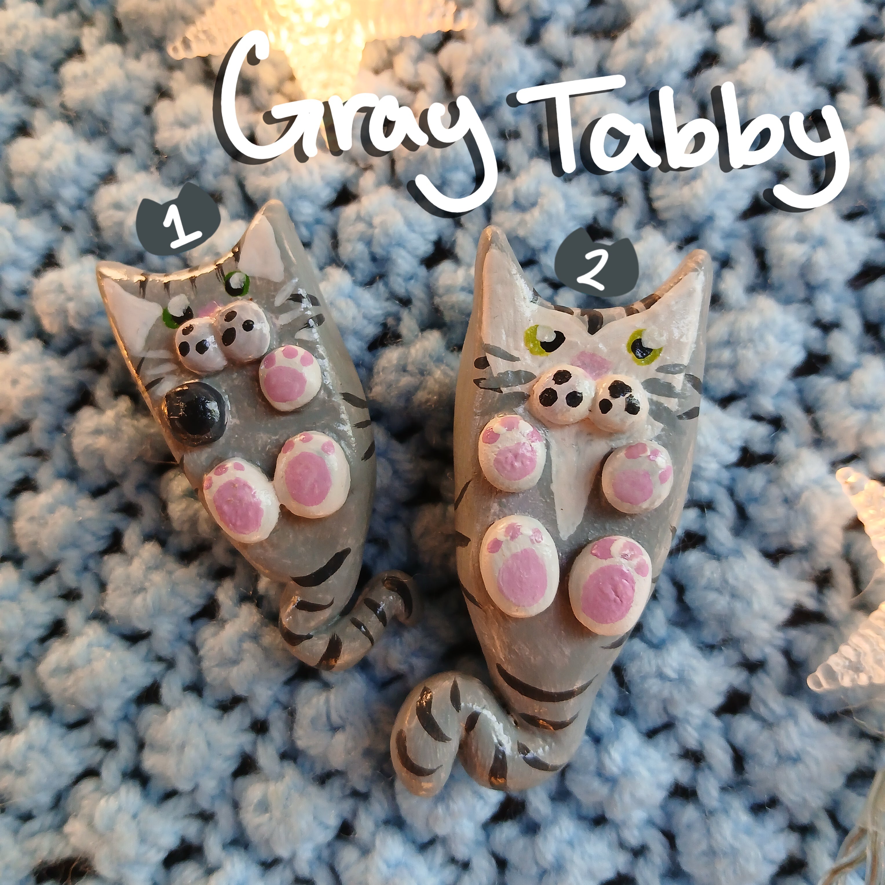 Two clay graytabby cat-shaped magnets, on light blue textured background, with decorative star lights.