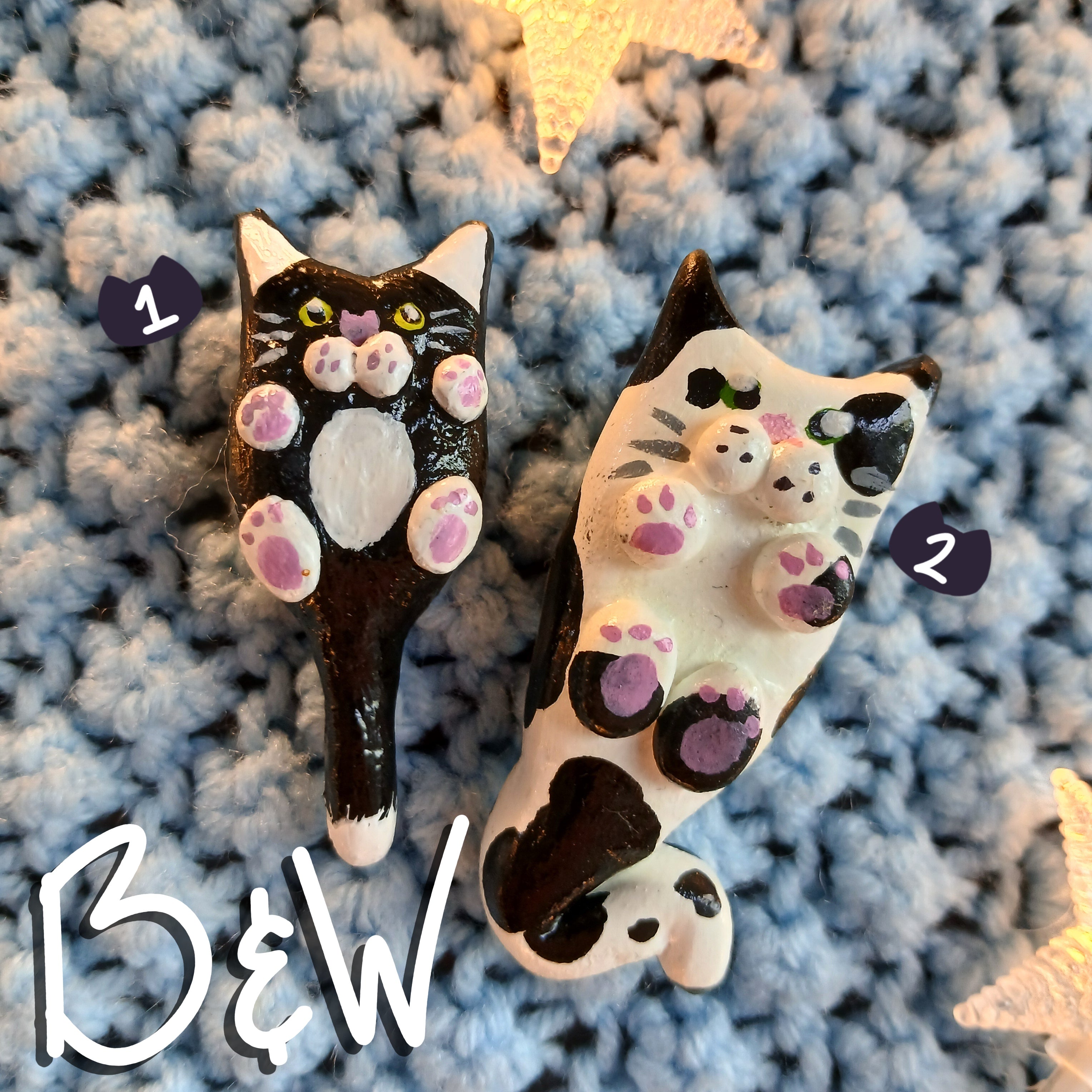 Two clay black and white cat-shaped magnets, on light blue textured background, with decorative star lights.